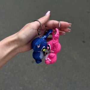 May include: Two crochet bunny keyrings, one blue and one pink, attached to a silver keyring. The blue bunny has a yellow star and the pink bunny has a heart. The keyrings are handmade and feature a cute, cartoonish design.