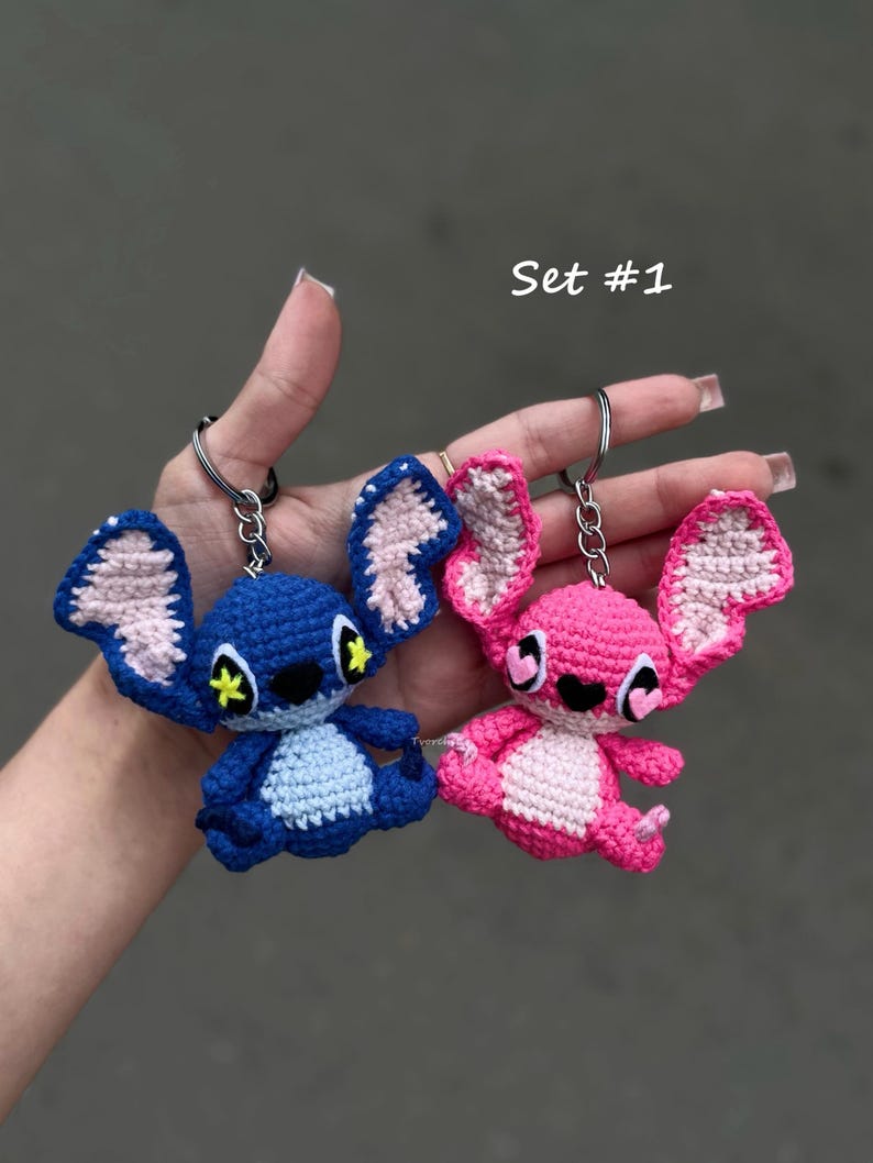 May include: Two crochet keyrings in the shape of cartoon characters. One is blue with pink details, the other is pink. Both have large ears, black eyes, and key rings. The text "Set #1" is visible.