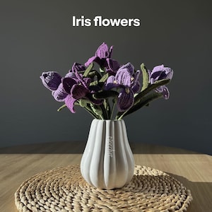 May include: A bouquet of crocheted iris flowers in a white vase, set on a woven mat. The flowers are a deep purple with green stems and leaves. The vase has a ribbed design and the text "for crochet" is visible.