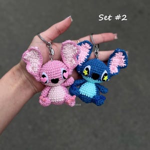 May include: Two crochet keyrings, one pink and one blue, resembling cartoon characters. The pink character has heart-shaped eyes and the blue character has yellow star accents. The text "Set #2" is visible.