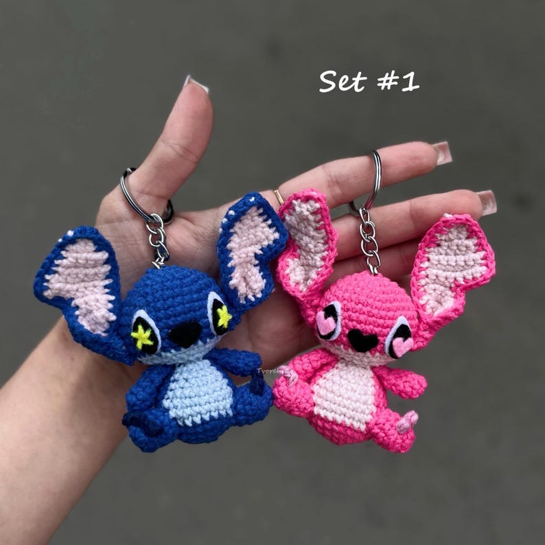May include: A set of two crochet keychains featuring cartoon characters. One is blue with yellow accents, the other is pink. Both have large ears and are attached to silver keyrings. The text "Set #1" is visible.