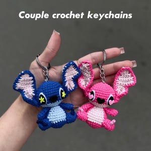 May include: Two crochet keychains, one blue and one pink, each with a cartoon character design. The blue keychain has yellow accents, while the pink one has pink heart accents. The text "Couple crochet keychains" is at the top.