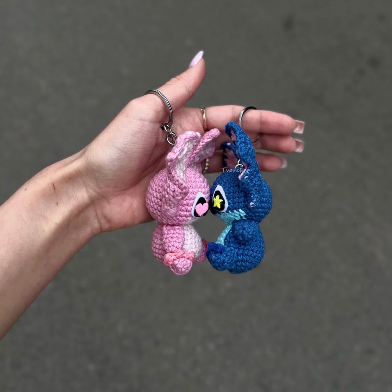 May include: Two crocheted keyrings, one pink and one blue, are held up against a grey background. The pink keyring has a heart-shaped face and the blue keyring has a star-shaped face. Both are in the shape of a bunny.