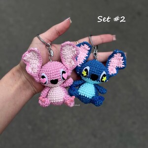 May include: Two crochet keychains, one pink and one blue, resembling a cartoon character. The pink keychain has pink ears and a white belly, while the blue one has blue ears and a light blue belly. Both have key rings attached and the text "Set #2".