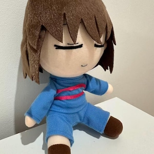 Puede incluir: Mu&ntilde;eco de peluche de un personaje con cabello casta&ntilde;o y ojos cerrados. El mu&ntilde;eco viste un atuendo azul con una raya rosa en el pecho y zapatos marrones. El mu&ntilde;eco est&aacute; sentado sobre una superficie blanca.
