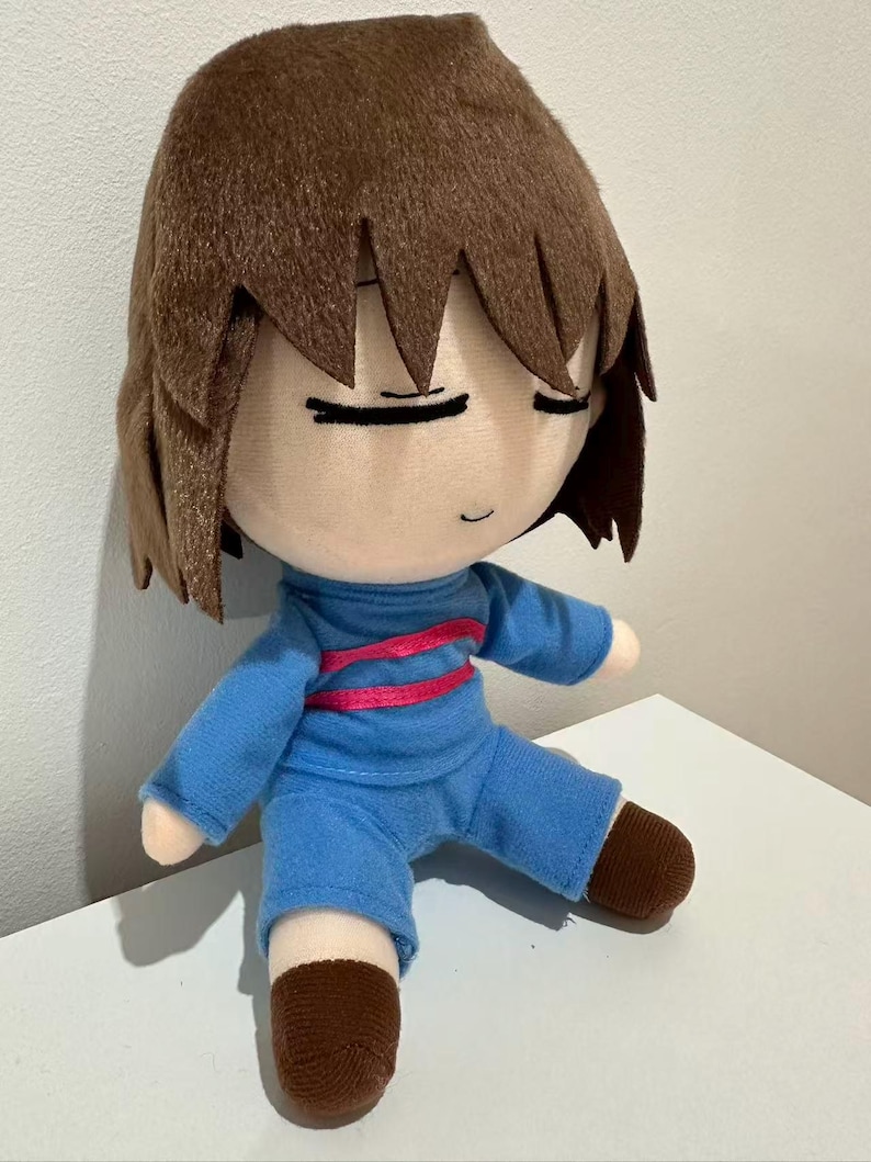 Puede incluir: Mu&ntilde;eco de peluche de un personaje con cabello casta&ntilde;o y ojos cerrados. El mu&ntilde;eco viste un atuendo azul con una raya rosa en el pecho y zapatos marrones. El mu&ntilde;eco est&aacute; sentado sobre una superficie blanca.