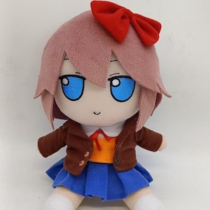 Puede incluir: Muñeca de peluche con un personaje de pelo rosa, ojos azules y un lazo rojo. La muñeca lleva una chaqueta marrón, una camisa naranja y una falda azul. Los calcetines y zapatos blancos completan el atuendo.
