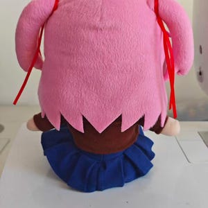 Puede incluir: Mu&ntilde;eca de peluche con pelo rosa en dos coletas laterales, cada una atada con una cinta roja. Lleva un top marr&oacute;n y una falda plisada azul. La mu&ntilde;eca est&aacute; sentada sobre una superficie blanca.
