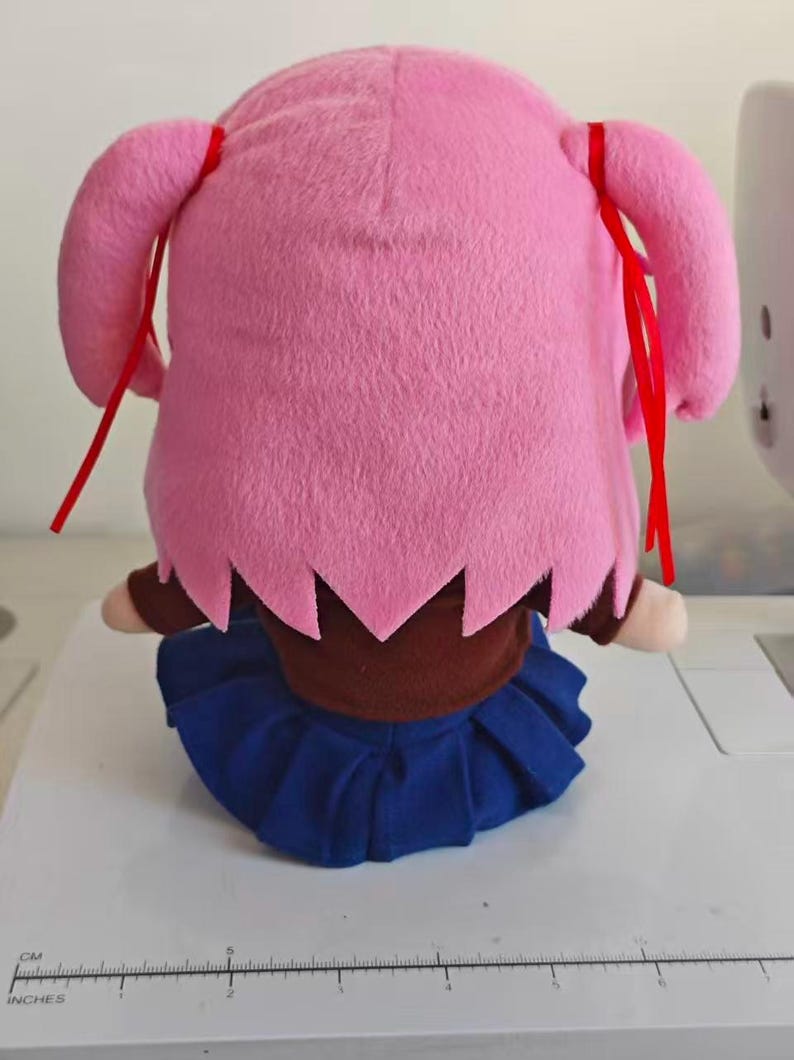 Puede incluir: Mu&ntilde;eca de peluche con pelo rosa en dos coletas laterales, cada una atada con una cinta roja. Lleva un top marr&oacute;n y una falda plisada azul. La mu&ntilde;eca est&aacute; sentada sobre una superficie blanca.