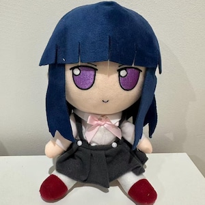 May include: A plush doll with a blue wig, purple eyes, and a small smile. The doll wears a white shirt with a pink bow, a gray skirt, and red shoes. The doll is sitting on a white surface.
