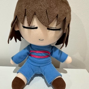 Puede incluir: Mu&ntilde;eco de peluche de un personaje con cabello casta&ntilde;o y ojos cerrados. El mu&ntilde;eco viste una camisa azul con una raya rosa y pantalones cortos azules. Los pies del mu&ntilde;eco son marrones.