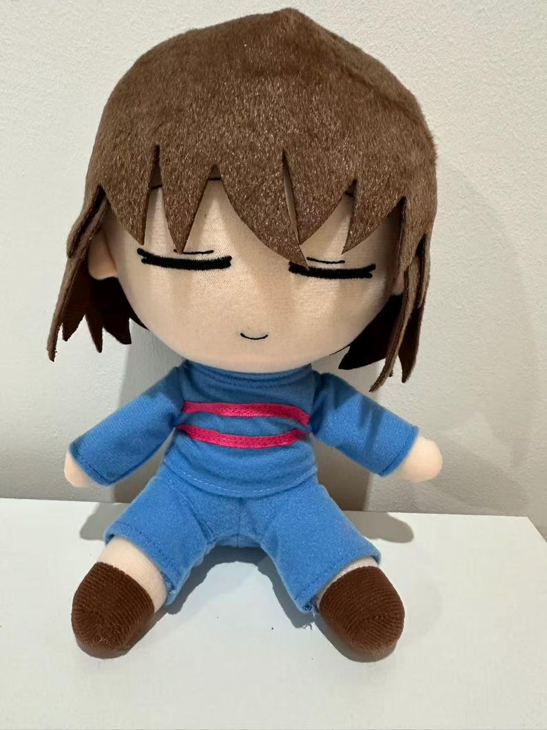 Puede incluir: Mu&ntilde;eco de peluche de un personaje con cabello casta&ntilde;o y ojos cerrados. El mu&ntilde;eco viste una camisa azul con una raya rosa y pantalones cortos azules. Los pies del mu&ntilde;eco son marrones.