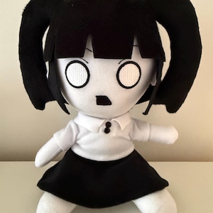 May include: A plush doll with a white face, black hair styled in two pigtails, and large circular white eyes. The doll wears a white collared shirt with two small black buttons and a black skirt. The limbs and face are made of white fabric.