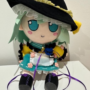 May include: A plush doll with mint green hair, large blue eyes, and pink cheeks, wearing a black hat with yellow trim, a yellow shirt, a teal apron with small pink roses, and black shoes with purple ribbons and heart-shaped accents.