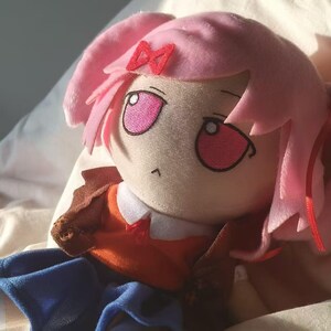 Puede incluir: Mu&ntilde;eca de peluche con pelo rosa y un lazo rojo. La mu&ntilde;eca tiene grandes ojos rosas y lleva una camisa naranja, una chaqueta marr&oacute;n y pantalones azules. La mu&ntilde;eca est&aacute; sobre una superficie blanca.