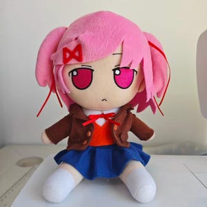 Puede incluir: Mu&ntilde;eca de peluche con un personaje de pelo rosa con coletas, chaqueta marr&oacute;n, falda azul y calcetines blancos. La mu&ntilde;eca tiene ojos grandes y un lazo rojo. La mu&ntilde;eca est&aacute; sentada sobre una superficie blanca.