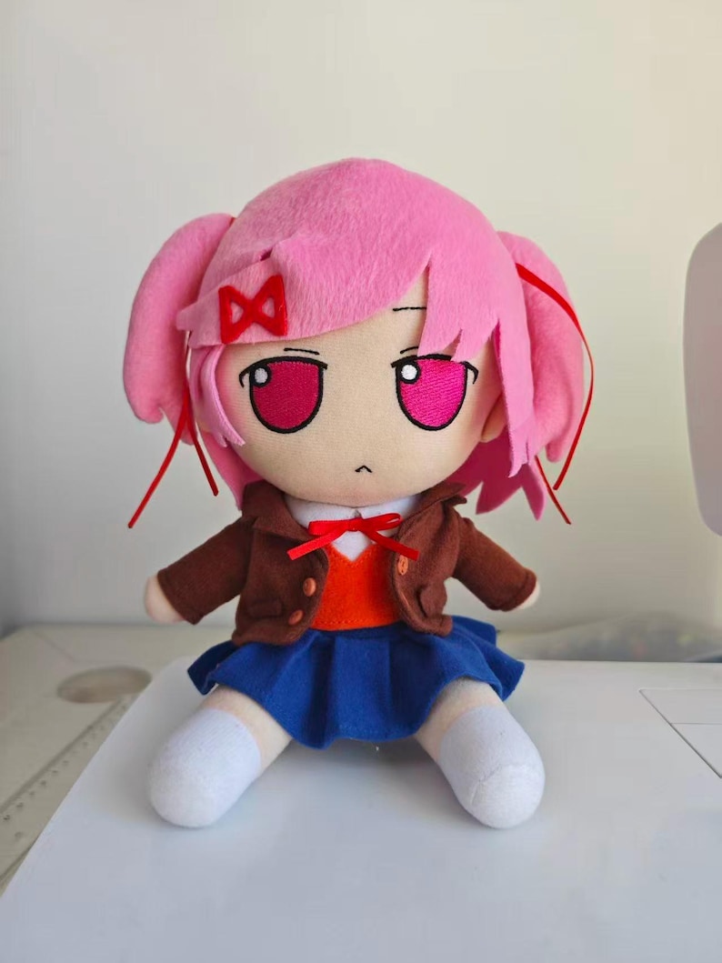 Puede incluir: Mu&ntilde;eca de peluche con un personaje de pelo rosa con coletas, chaqueta marr&oacute;n, falda azul y calcetines blancos. La mu&ntilde;eca tiene ojos grandes y un lazo rojo. La mu&ntilde;eca est&aacute; sentada sobre una superficie blanca.