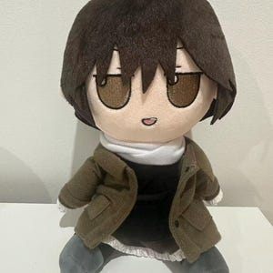 May include: A plush doll featuring a character with short dark brown hair, brown eyes, and a small mouth. The doll wears a dark olive green jacket over a black dress with a white collar and white trim at the hem, paired with dark grey shoes.