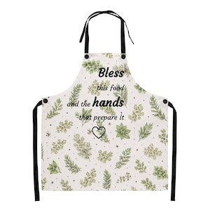 May include: White apron with black straps and ties, decorated with a botanical pattern of green leaves and flowers. The apron has the text "Bless this food and the hands that prepare it" with a heart symbol.