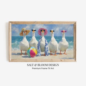 Puede incluir: Una pintura enmarcada representa cinco gansos blancos en una playa, cada uno con gafas de sol y atuendo playero. Un ganso lleva un sombrero de paja, otro un sombrero rosa y un tercero una envoltura a rayas azules y blancas. Una colorida pelota de playa descansa sobre la arena. La obra de arte está etiquetada como "SALT & BLOOM DESIGN."