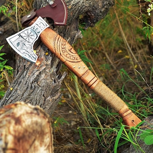 May include: A detailed axe with a wooden handle and a metal blade. The handle is carved with a design, and the blade has an engraving. A brown leather sheath protects the axe head. The axe is resting on a tree trunk, surrounded by greenery.