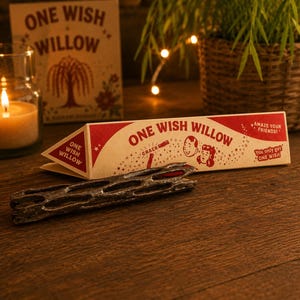 May include: A triangular cardboard box labeled One Wish Willow with red text and illustrations of two faces, featuring the phrases Amaze Your Friends and You only get ONE WISH. A dark, porous, cylindrical wooden stick lies in front of the box.