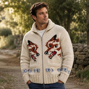 May include: A cream-coloured, chunky knit zip-up cardigan featuring a shawl collar and two front pockets. The sweater displays a repeating pattern of two fox faces in brown, black, and white, with a blue floral-style border along the bottom hem.