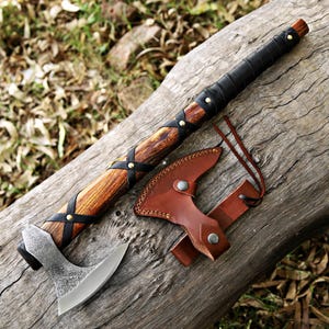 Peut inclure: Une hache artisanale avec un manche en bois enveloppé de cuir noir et fixé avec des rivets en laiton. La tête de la hache est de couleur argentée et possède une lame incurvée. Un étui en cuir marron est inclus.