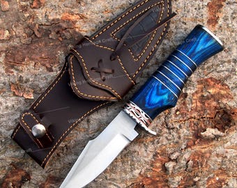 May include: A hunting knife with a polished steel blade and a blue wooden handle. The handle is accented with silver rings and a decorative guard. The knife is paired with a brown leather sheath with decorative stitching.