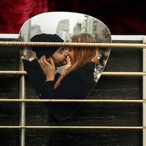 May include: A custom guitar pick featuring a printed photograph of a couple embracing, positioned behind the metallic strings of a guitar. The pick has a smooth, rounded triangular shape and displays a clear, high-resolution image of two people.