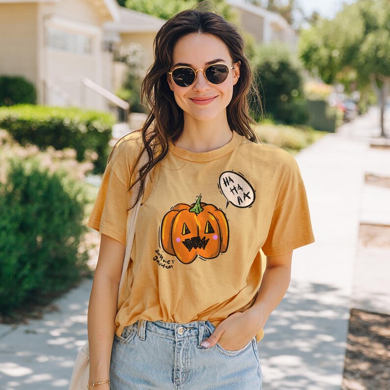 May include: Mustard yellow t-shirt featuring a cartoon pumpkin with a smiling face and the text "HA HA HA" in a speech bubble. The shirt also has the text "Annie James" on the bottom left.
