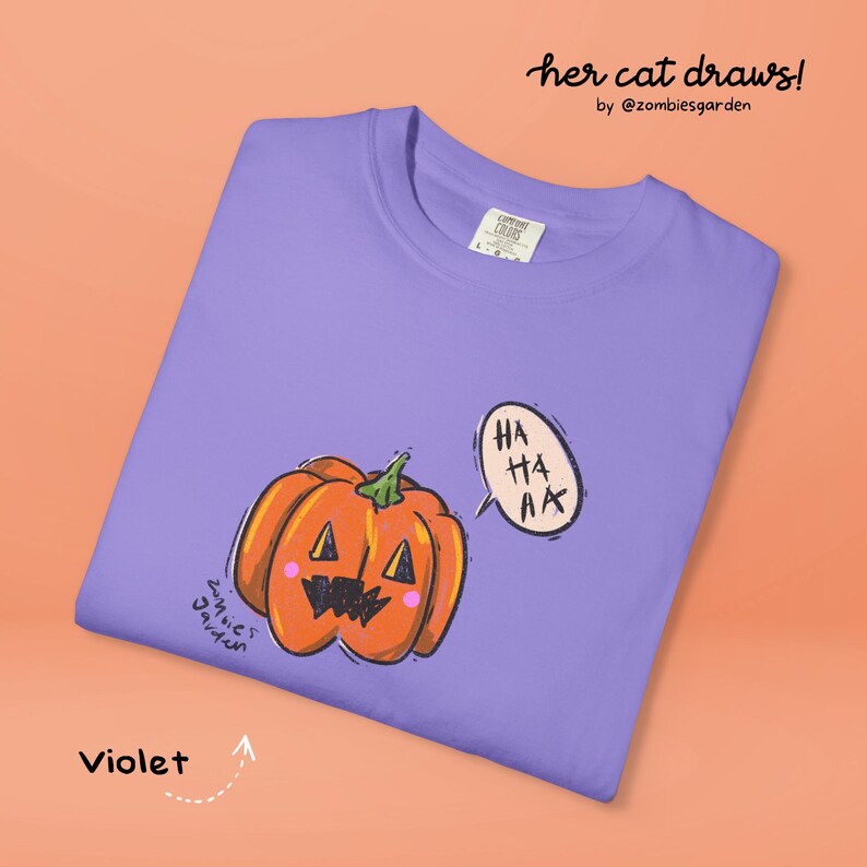 May include: Violet t-shirt featuring a cartoon pumpkin with a smiling face and a speech bubble that says "HA HA AA". The shirt has the text "her cat draws!" and "by @zombiesgarden".