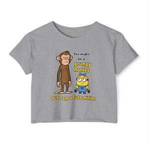 May include: A gray t-shirt featuring a grumpy monkey and a cute minion graphic. The text reads "You might be a grumpy monkey but I am a cute minion". The shirt is made of a soft material.