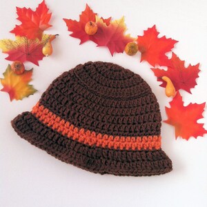 May include: A handmade brown crochet hat with an orange stripe around the crown. The hat is surrounded by fall-themed decorations, including red and yellow artificial leaves and small gourds. The hat has a brim and a rounded top.