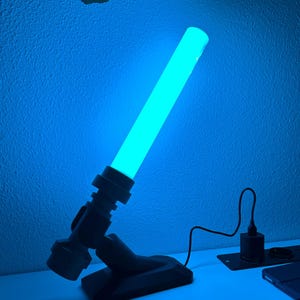 May include: A blue glowing lightsaber-style lamp. The lamp is angled, with a cylindrical light source and a black base. A power cord and charging base are also visible.