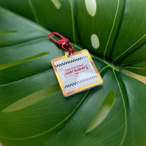 May include: A clear acrylic keyring with a red clasp. The keyring features the text "Fresh Oven Baked! FREDDY FAZBEAR'S Pizza" on a white background with a black and white checkered border and a gold trim.