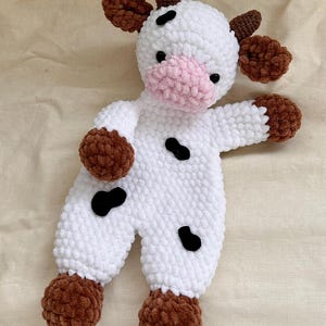 May include: A handmade crochet cow plush toy. The toy is primarily white with brown accents on the ears, hooves, and horns. It has a pink muzzle and black spots. The plush is soft and cuddly.