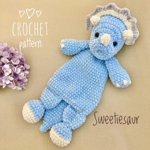 Op de afbeelding: Een lichtblauw gehaakt triceratops knuffel met witte en grijze accenten. Het speelgoed heeft zwarte ogen en het woord "Sweetiesaur" staat op de zijkant. De woorden "CROCHET pattern" zijn ook zichtbaar.