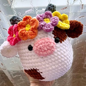 May include: A handmade, white crocheted cow head with brown ears and a pink nose. The cow is adorned with a colorful flower crown featuring orange, pink, yellow, and purple flowers. Black button eyes complete the whimsical design.