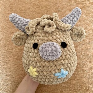 May include: A crocheted cow head in tan, grey and black. The cow features grey horns, ears, and a nose, with black button eyes. Two small flower accents in yellow and blue are on the side of the face.