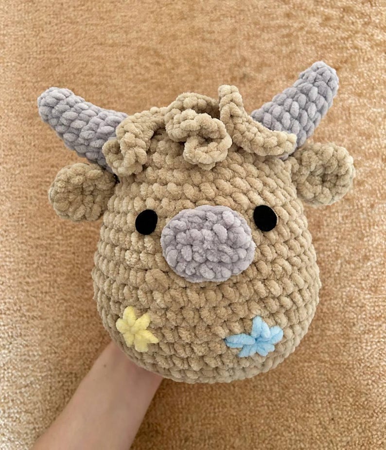 May include: A crocheted cow head in tan, grey and black. The cow features grey horns, ears, and a nose, with black button eyes. Two small flower accents in yellow and blue are on the side of the face.