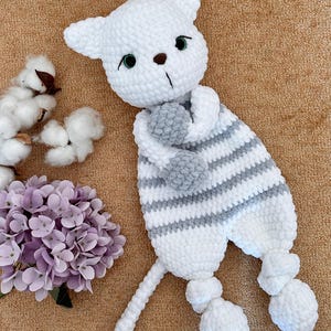 Puede incluir: Un juguete de gato de ganchillo a rayas blancas y grises hecho a mano, con ojos verdes y una nariz marrón. El gato tiene una cola larga y dos pies redondos. El juguete está sobre una superficie marrón junto a un ramo de flores moradas y algodón.