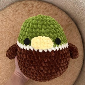 May include: A handmade, plush duck toy. It has a green head, brown body, and small brown wings. The duck features a yellow beak, black eyes, and a white trim around the neck. The toy is made of soft, textured yarn.