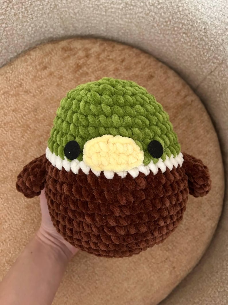 May include: A handmade, plush duck toy. It has a green head, brown body, and small brown wings. The duck features a yellow beak, black eyes, and a white trim around the neck. The toy is made of soft, textured yarn.