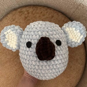 May include: A handmade, light blue crochet koala head with a brown nose and black eyes. The koala has fluffy ears with white and light blue accents. This soft, plush toy is perfect for children.
