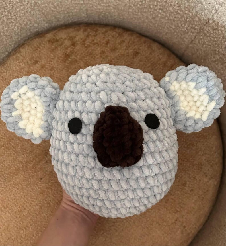 May include: A handmade, light blue crochet koala head with a brown nose and black eyes. The koala has fluffy ears with white and light blue accents. This soft, plush toy is perfect for children.