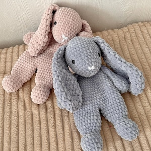 Peut inclure: Deux jouets en forme de lapin en peluche faits à la main. L'un est rose clair, l'autre gris. Ils ont de longues oreilles tombantes, de petits yeux noirs et des détails blancs. Posés sur une surface texturée de couleur beige.