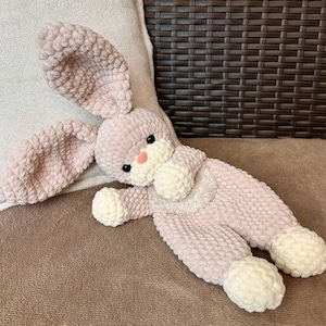 May include: A handmade plush bunny in soft pink with white accents. The bunny has long, floppy ears, black eyes, and a pink nose. Its body is elongated, with white paws and a fluffy, textured appearance. The toy is lying on a brown surface.