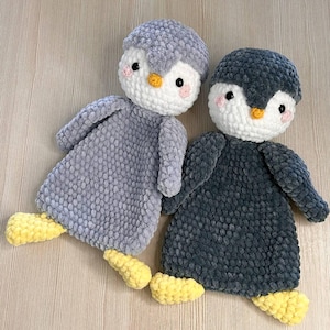 May include: Two plush penguin toys. One is gray, the other is dark gray. Both have white faces, yellow beaks and feet, and pink cheeks. The penguins are lying on a light brown surface.