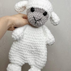 May include: A handmade white crochet lamb stuffed animal with a gray face and black eyes. The lamb has a fluffy texture and is designed with small ears, arms, and legs. The lamb is a soft toy.